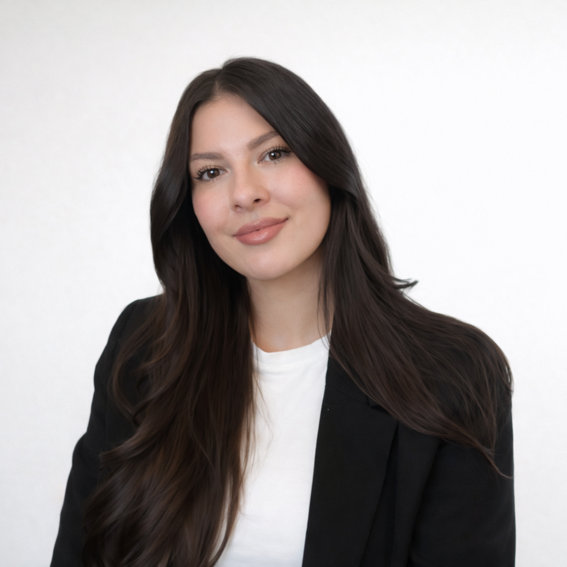 Eine Frau mit langen dunklen Haaren, die ein weißes Hemd und einen schwarzen Blazer trägt, lächelt sanft, während sie vor einem schlichten hellen Hintergrund sitzt.