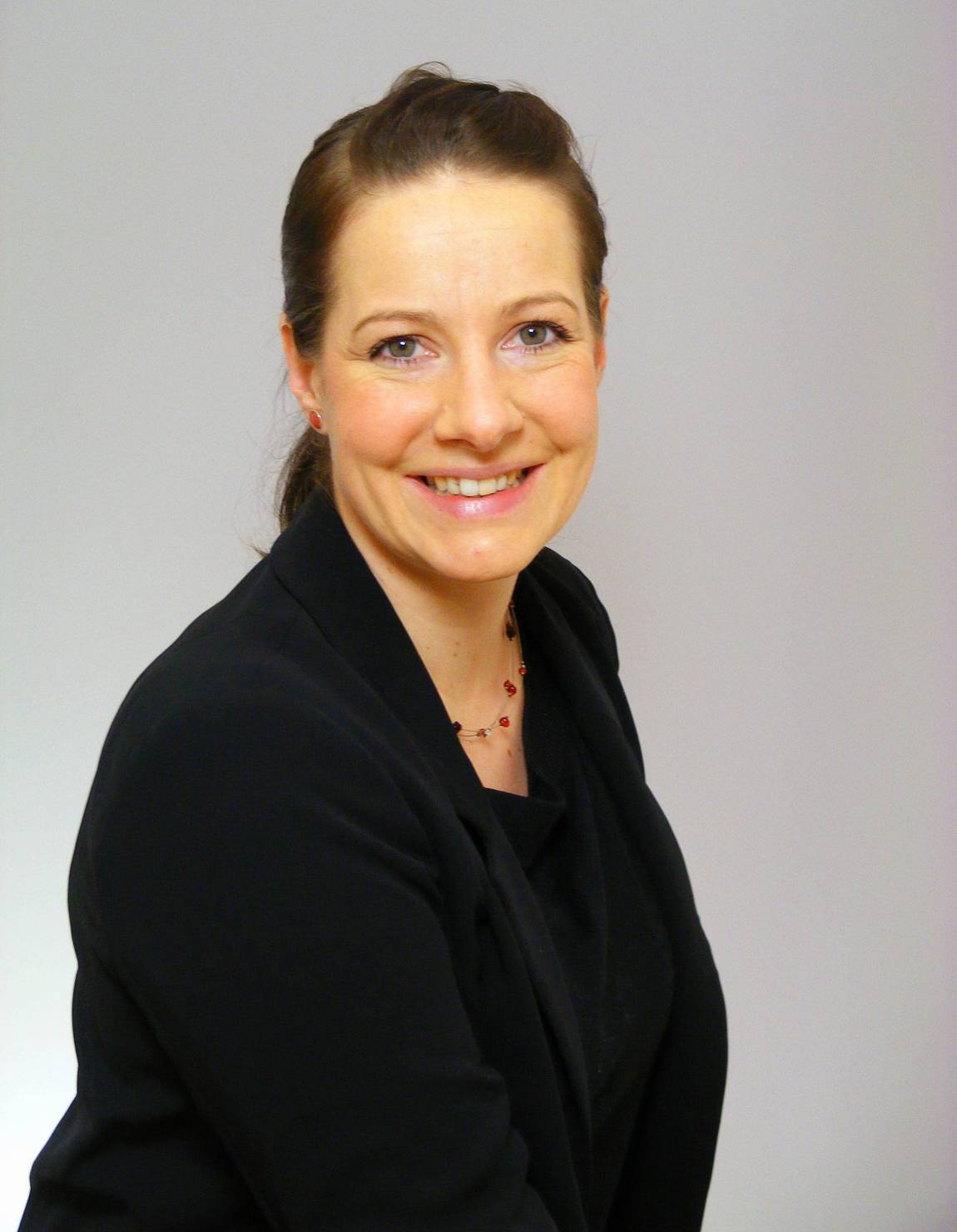 Eine Frau mit heller Haut und zurückgebundenem braunem Haar, die einen schwarzen Blazer und eine zarte Halskette trägt, lächelt in die Kamera vor einem schlichten hellen Hintergrund.